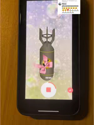 A smartphone screen displays a stylized bomb with a large pink bow, set against a blurred, bokeh-filled background. The phone rests on a wooden surface. The interface shows a recording progress indicator.