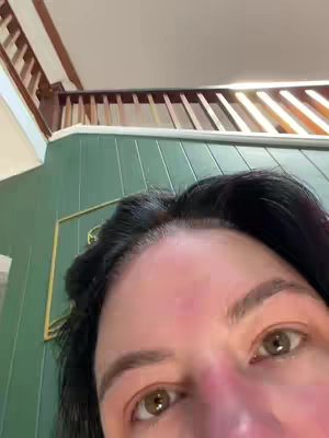 A close-up shot from the viewer's perspective, focusing on part of a person's forehead and eyes. The eyes, colored a light brown, are the center of attention. The person has dark hair; some of the hair covers the top of the image. The setting is indoors, with a light green wall visible behind the person.