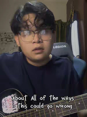 A young man wearing glasses is centered in a video frame, likely indoors. He is positioned in a medium shot, with his upper body visible. His expression suggests contemplation or introspection. A gray gaming chair with the word "LEGION" is visible behind him. The setting appears to be casual, possibly a bedroom based on the soft curtain in the background, and a partially visible wardrobe. A guitar is held in front of him, with overlay text on the guitar that reads the lyrics of the song being sung.