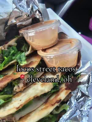 The image showcases a close-up of a pile of tacos, with fresh ingredients and a side of dipping sauce in small plastic containers. The tacos are sitting on a piece of shiny aluminum foil, with the image shot at a slight angle.