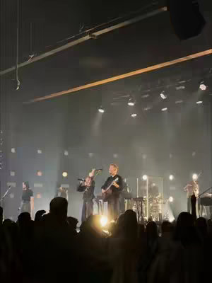 A live music performance on stage with band members, a crowd is visible. The stage is lit with stage lighting.