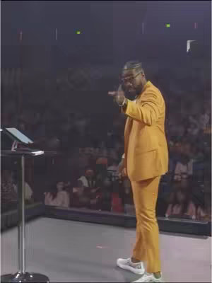 A man in a mustard-colored suit and white sneakers speaks on a stage, gesturing with his right hand. He stands beside a podium with a tablet, with a dark background suggesting an audience.
