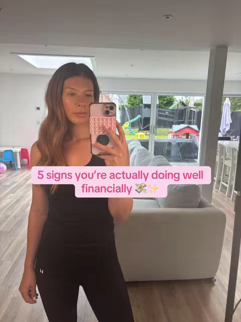 A woman with long brown hair is taking a selfie with her phone in front of a mirror. She's wearing a black tank top and leggings. Overlaying the image is text that says '5 signs you're actually doing well financially'. The background includes a living room with a large window and a view of a backyard.