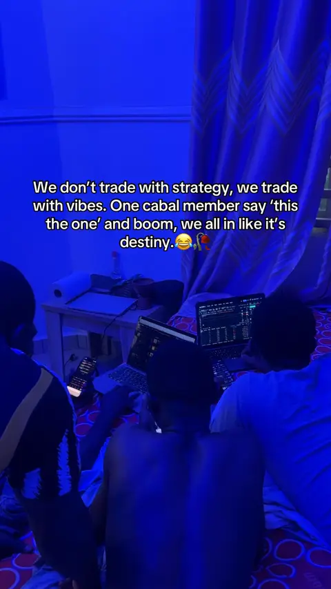 The image shows a dimly lit room with blue lighting. Three men are partially visible, gathered around a laptop computer and a smartphone, suggesting a group activity. Text overlayed on the image includes a quote about trading with 'vibes' rather than strategy, implying a focus on financial markets or trading strategies. A curved curtain with a repeating pattern is visible in the background.