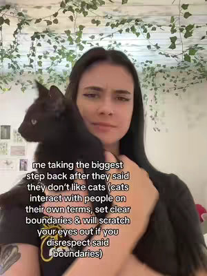 A young woman with dark hair and a black top holds a black cat close to her face, with text overlayed on the image. The background includes a wall with vines and some photos hung on it.