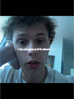 A young man with short hair, wearing a white t-shirt and a silver chain, is looking directly at the camera with a serious expression. Text overlaid in a stylized font at the center reads: "I do not give a f*k about girls." The background is blurred, suggesting an indoor setting.