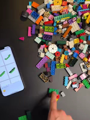 A top-down shot shows a black surface covered in a colorful assortment of LEGO bricks. An iPhone displaying a screen from the 'Brickit' app rests to the left, while a hand with a finger extended touches a cluster of bricks in the bottom right. The composition is casual and inviting.
