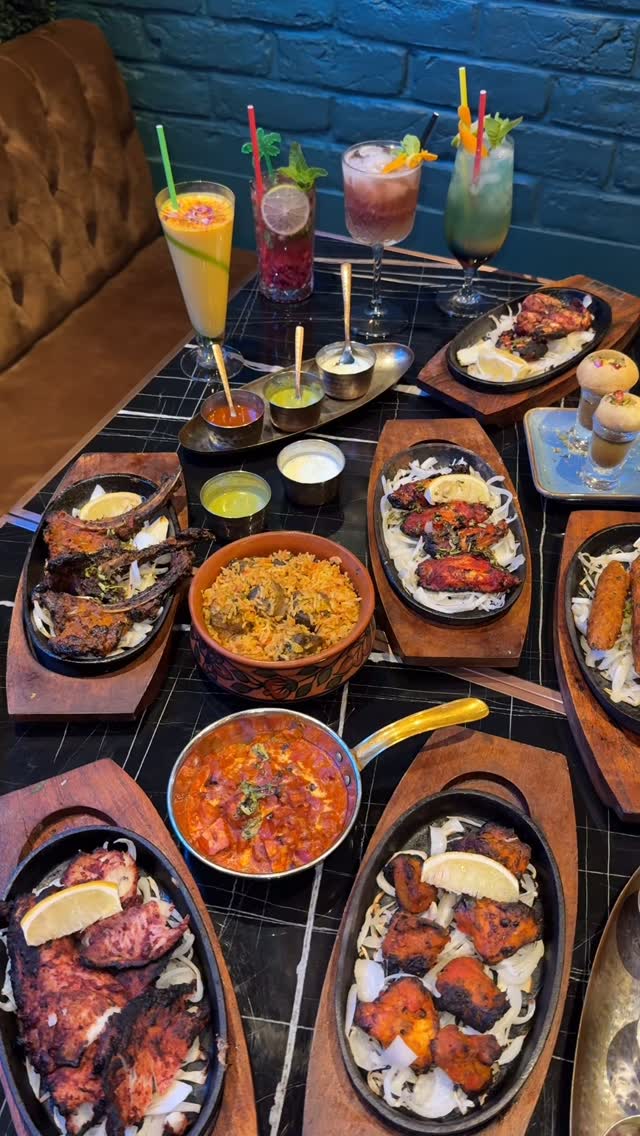 An overhead shot of a table laden with Indian food and drinks. Several dishes are displayed in black oval-shaped skillets and bowls, including lamb chops, chicken wings, and a bowl of biryani. Cocktails are arranged at the top, along with small containers of sauces. The table surface has a black and white checkered pattern, and the background shows a hint of a dark wall.