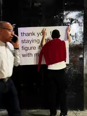A photo taken at eye-level, in an outdoor setting. A person is holding a sign with their back to the viewer while a second person, in the left of the frame, appears to be speaking on a phone. The person writing the message wears a red sweater draped over their shoulders, a white t-shirt, and black pants.
Behind the person, is a dark, textured wall. The second person wears glasses and a white long-sleeved shirt.