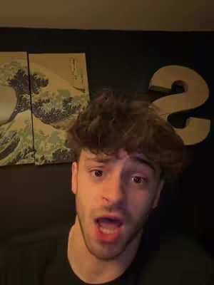 A young man is looking directly at the camera with a surprised expression, mouth open. Behind him are two framed art prints depicting a wave from a Japanese artwork, and a large, three-dimensional number "2" is placed to the right of the man's head against a black wall.