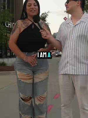 A woman with tattoos and distressed jeans is being interviewed on a city street by a man in sunglasses. A text overlay reads 'I AM A'.