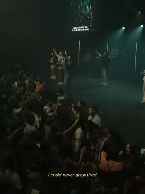 An image capturing a lively worship service. A large crowd faces a stage where individuals are singing and raising their hands. The stage is lit with spotlights and a large screen displays text. The overall color scheme is dark with bright lights highlighting the performers and the crowd, conveying a sense of energy and spiritual engagement.