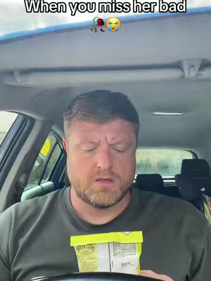 A man with a beard sits in a car, holding a snack bag, with a concerned expression.
