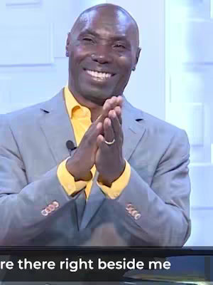 The image shows a middle-aged African-American man in a studio setting. He is bald, wearing a yellow dress shirt under a gray blazer, and has a gold ring on his left hand. He is smiling with his hands clasped in front of his chest. The background is a light-colored, repeating pattern. At the bottom of the image shows the start of some text overlaid.