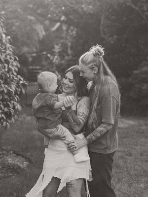 A grayscale photo features a family of three: two women and a child. The women, presumably the mothers, embrace the child, creating a warm and intimate scene. The background is blurred, suggesting an outdoor setting, possibly a garden, with soft, natural lighting enhancing the tender moment between the parents and their child.