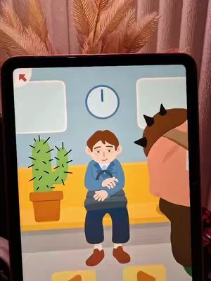 An iPad screen displays a cartoon scene of a man sitting on a bench in what appears to be a subway or bus station. A large cactus is to the left, and another figure with a spiky head is visible on the right. The scene is rendered in a simple, colorful style, and the overall composition is well-lit and appealing.