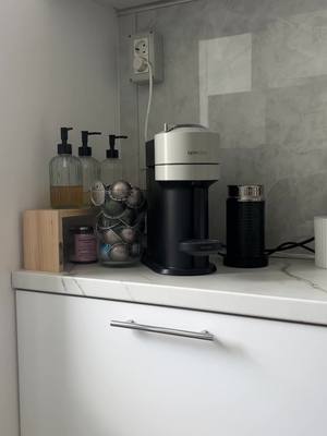 A photo of a kitchen counter with a coffee machine, espresso pods, and accessories like soap dispensers and a milk frother. The setting is neat and modern. The wall is white and has a marble-like pattern.
