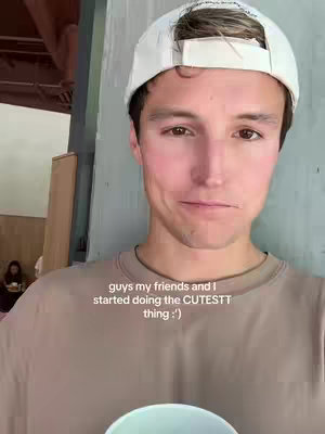 A young man in a baseball cap smiles, looking down at what appears to be a cup, in front of a light green wall. A woman is blurred in the background sitting in a cafe.