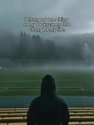 The image shows a person in a hooded sweatshirt standing on what appears to be stadium seating, facing a sports field under a cloudy sky. The person is silhouetted, with the back of their head and shoulders visible. Above, text overlays the image. The overall tonality is dark and atmospheric.