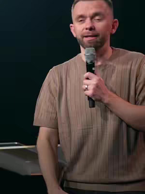 A medium shot of a man standing in front of a dark background, holding a microphone. He is wearing a light brown, short-sleeved shirt with a subtle ribbed texture. The man has short hair and a slight smile. His right hand holds the microphone, and a ring is visible on his left ring finger. In the background, a portion of a dark surface is visible, possibly a desk or podium.