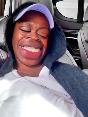 A close-up shot of a person inside a car. The person is wearing a white shirt and a hoodie with the hood up, as well as a purple cap. Their face is distorted, with their eyes closed and mouth wide open in a exaggerated, crying-like expression. The overall effect is comedic.
