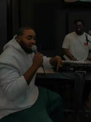 The cover image shows two Black men in a recording studio setting. The main subject is seated and singing into a microphone, wearing a white hoodie and green pants. A second person, wearing a white t-shirt, plays a keyboard in the background. The lighting is dim, creating a somber atmosphere enhanced by dark setting.