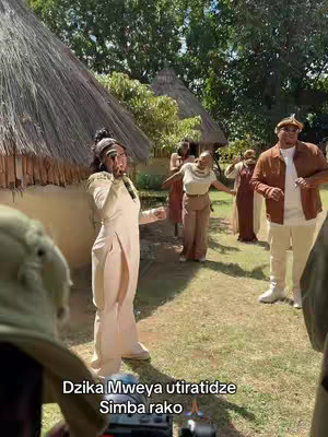 The cover image captures a lively scene outdoors, featuring a woman in a cream-colored dress as the central figure. She appears to be singing, with arms outstretched. Surrounding her are other people in various costumes, suggesting a performance or celebration. The setting includes a traditional African village style with thatched roof buildings and green grass. The overall composition is bright and full of movement, likely indicative of a cultural or musical celebration.