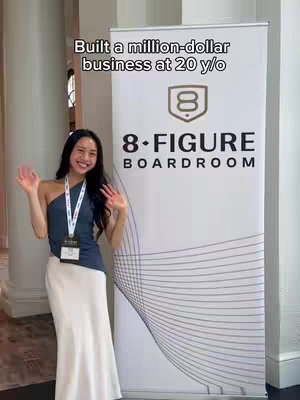 A woman in a formal setting with a banner behind her promoting '8-FIGURE BOARDROOM', the text hook above says 'Built a million-dollar business at 20 y/o'. She is waving at the camera, smiling. The background is a hallway.