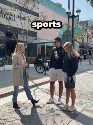 The image captures a street scene with three people interacting: a woman wearing a light-colored coat and jeans standing on the left, and two people to her right, one holding a cup and wearing sunglasses. The word "sports" is overlaid on the image. The background shows an outdoor environment with buildings, trees, and a bike.