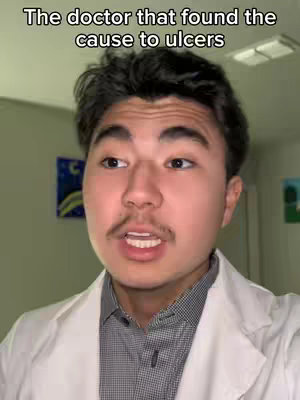 A man wearing a white lab coat speaks with a serious expression, set against the backdrop of a plain wall with framed pictures.