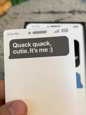 A person's hand is holding a white card with a text bubble on it. The screen of a cell phone is visible in the background.