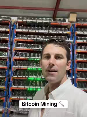 A man is standing in front of racks packed with computer servers or mining equipment. The man is wearing a light-colored shirt and speaking. The background is a large warehouse style space with industrial lighting.
