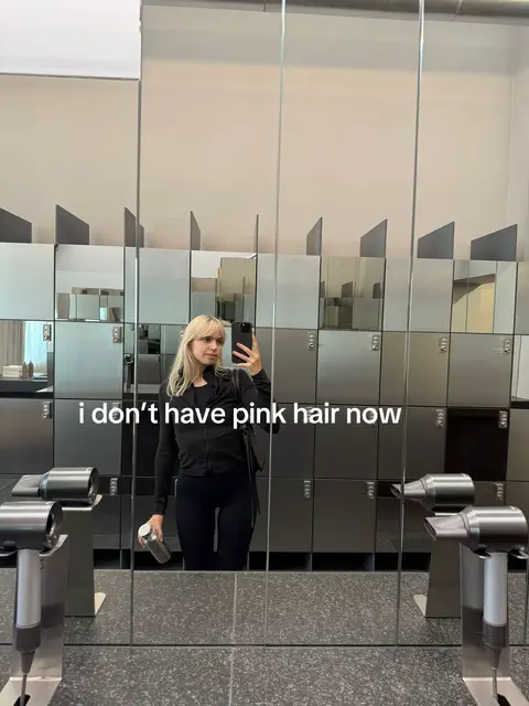 A woman is taking a mirror selfie in what appears to be a locker room or gym changing area. Behind her, mirrored lockers create a repeating pattern of reflections, and there are two hair dryers visible, adding a functional context to the space. The woman is wearing a black outfit.