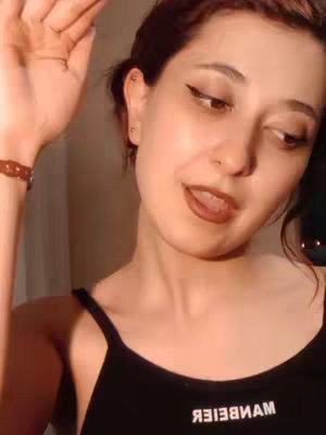 A selfie-style shot with a woman looking towards the right, her mouth slightly open, and her right hand raised. She wears a black tank top with a word printed in reverse, dark eyeliner, and brown lipstick. The background is a neutral color, likely a wall.