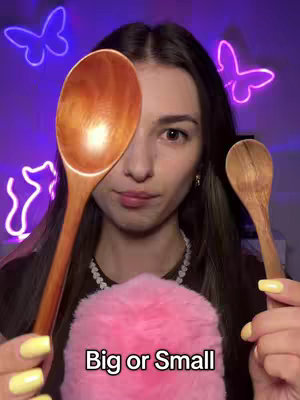 A woman holds a large wooden spoon in front of her left eye, with a smaller spoon in her right hand. A pink fluffy object sits below her face, and the background is a purple wall illuminated with neon butterfly and cat silhouettes. The image is well-lit and focuses on the woman's face and the objects she is holding.