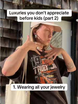A person with blonde hair is facing away from the camera, raising both hands with jewelry on their fingers, and wearing a baseball cap and T-shirt. Text overlays read "Luxuries you don't appreciate before kids (part 2)" and  "1. Wearing all your jewelry"
