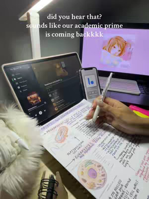 The cover image shows a well-lit desk setup for studying. A laptop is open, displaying what appears to be a podcast or music app, and a smartphone is propped up beside it. A biology study sheet with colorful diagrams is spread out in front. A hand holding a pen is positioned over the notes, ready to write, with an additional screen showing a cartoon on the background. There is also a furry white cushion or rug. The overall feel is focused on studying.