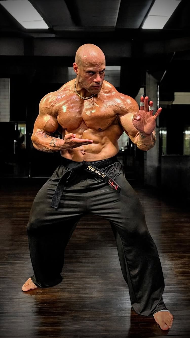 A muscular man in a karate stance is the central focus, set against a dark gym backdrop. He poses with one hand open and the other in a fist, wearing a black belt and pants. The lighting is dramatic, highlighting his physique with a dark background.