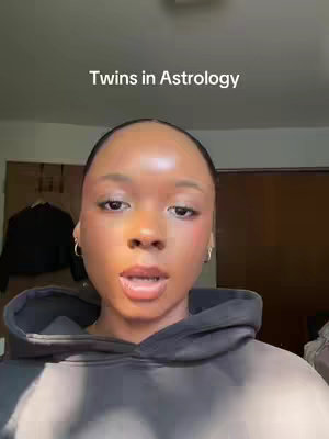 A close-up shot of a woman with brown skin and dark hair pulled back, wearing a gray hoodie. She is centered in the frame against a neutral background. The lighting is soft, highlighting her features. Text overlay at the top reads "Twins in Astrology".