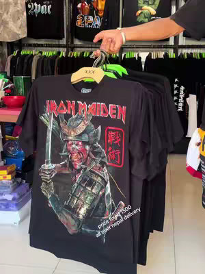 A person holds up a black t-shirt featuring an Iron Maiden band graphic in a shop setting. The shirt is printed with an image of the band's mascot, Eddie, in a samurai-inspired design holding a sword, with red text, and a red seal-like element with Japanese characters. The background shows racks of shirts, indicating a clothing store. There are some signs and decorations visible in the background.