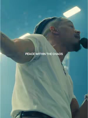 A man with dark skin, wearing a white t-shirt, is singing into a microphone with the text 'PEACE WITHIN THE CHAOS' overlaid on the image. The lighting is bright and the background is blurred, suggesting a performance setting.