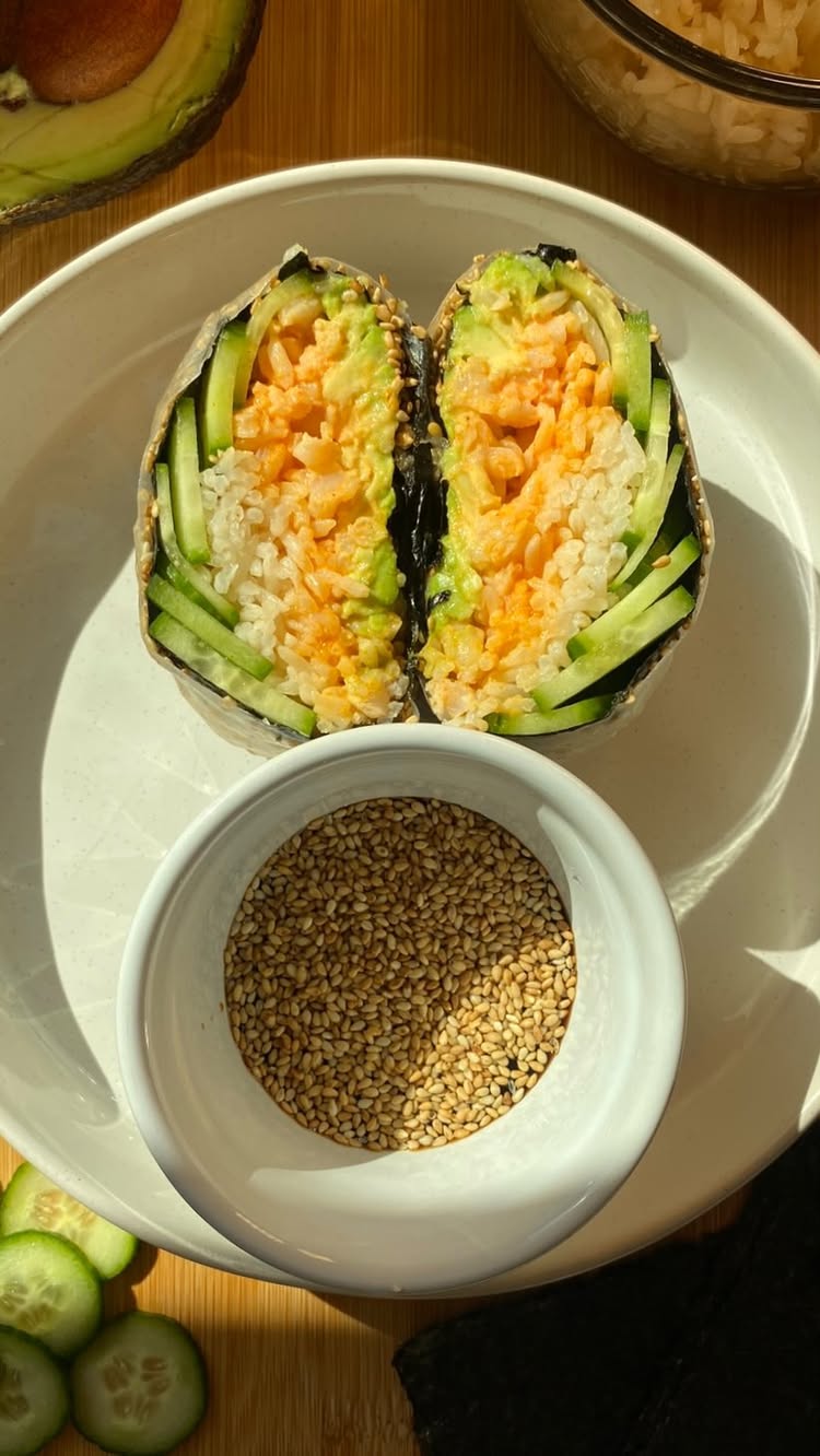 The image features a close-up of a spicy tuna sandwich, cut in half to showcase its colorful layers. The sandwich is wrapped in nori seaweed, filled with white rice, spicy tuna mix, and sliced cucumber and avocado. A small white bowl filled with sesame seeds sits in front of the sandwich, and a partial avocado is seen at the top.