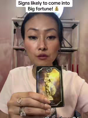 A woman with dark hair and wearing a light pink shirt stands in front of a neutral background. She holds a tarot card in front of her, with a focused expression. The card shows a woman with a yellow background.  Text overlays appear at the top of the frame.