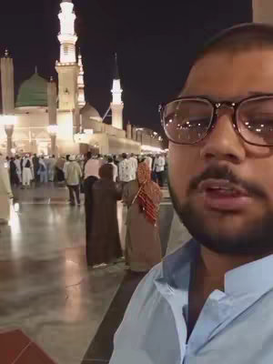 A man with glasses stands in front of a brightly lit mosque at night. The camera is close to the man's face, placing him in the foreground.