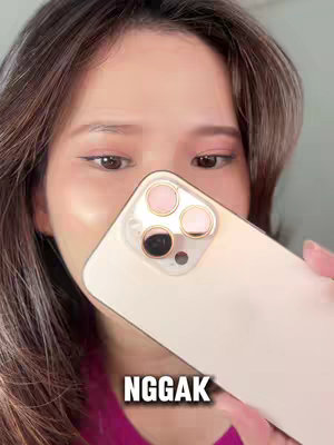 A woman's face is partially obscured by a gold iPhone. The upper half of her face is visible, showing her eyes and forehead with makeup. The screen displays the phone’s camera configuration. The background is a light color.