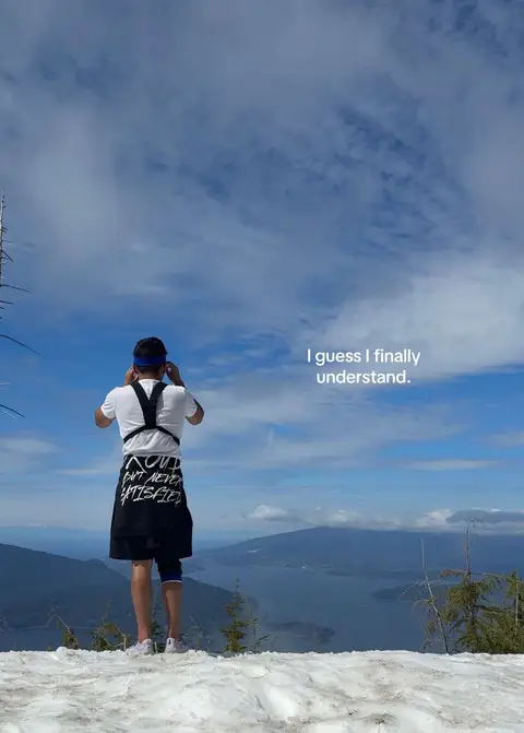 A man stands on a snowy mountaintop, viewed from the back. He is wearing a white shirt, black shorts, and a blue headband. The background is a vast display of blue sky and white clouds, with a scenic view of a body of water and mountains in the distance.