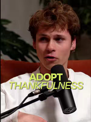 A young man with fair skin and short, wavy brown hair is centered in the frame, looking slightly to the side and speaking into a black microphone. The background is out of focus, with a hint of orange and green suggesting a room setting. Overlayed on the image is bright yellow text.