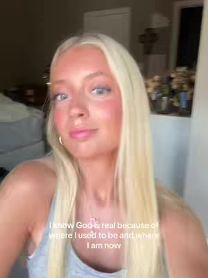 A young woman with long blonde hair and a pink blush poses for a selfie. She is wearing a gray tank top and a necklace. Text on the screen says, "I know God is real because of where I used to be and where I am now".
