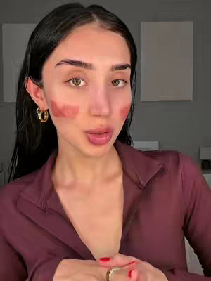 A woman with dark hair and fair skin is centered in the frame, looking at the camera with a slight smile. Patches of reddish makeup are on both of her cheeks. She is wearing a maroon-colored collared shirt and gold hoop earrings. The background has two white rectangular shapes, and a gray wall.
