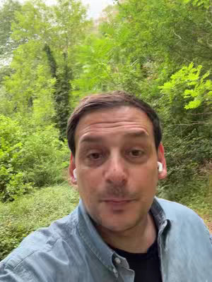 A man with brown hair and a blue shirt stands in front of a blurry green background of trees and foliage. The man is looking at the camera and wearing white ear buds.